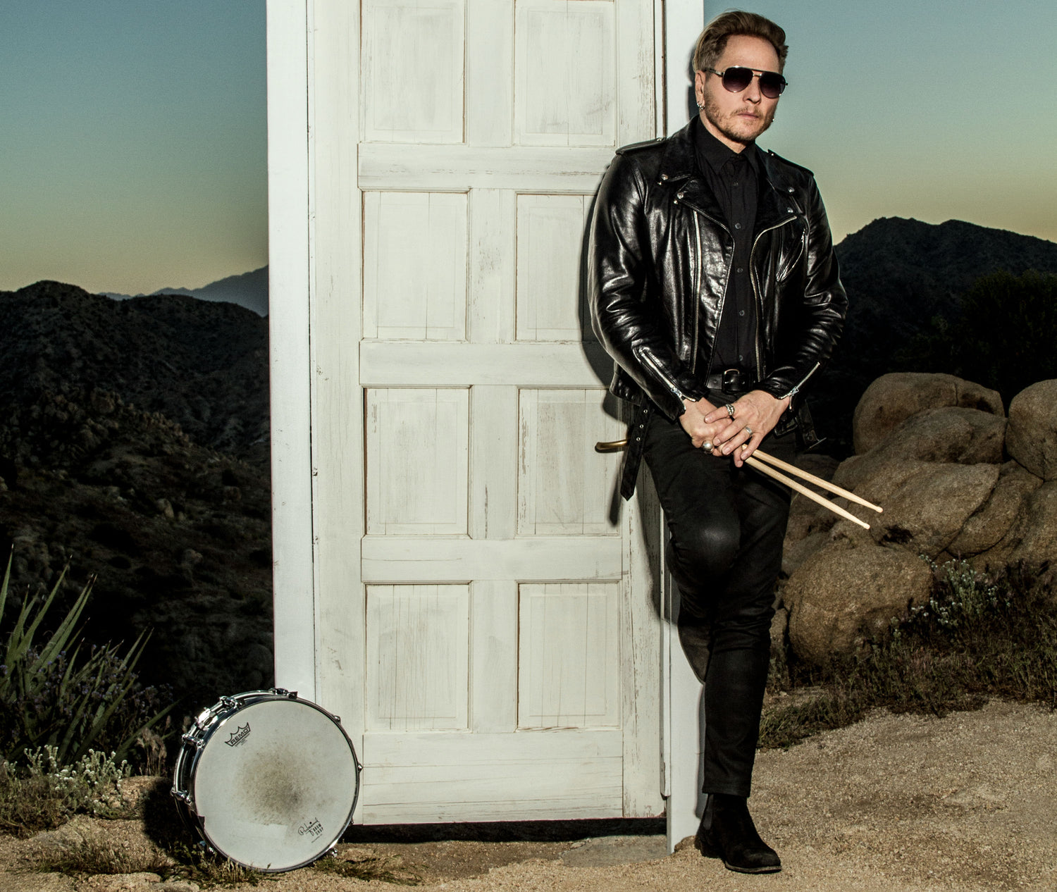 Matt Sorum Portrait for The Art of Giving standing against doorway in desert drumsticks in hand snare drum set against door frame