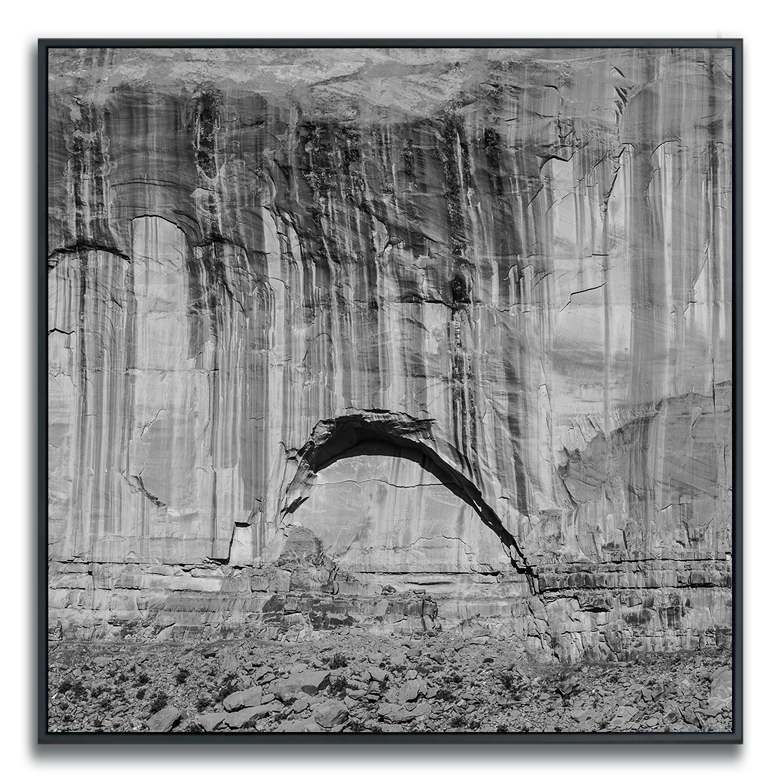 Black-and-white photograph of a dramatic rock formation with a natural arch spanning a rugged cliffside. The image highlights the textures and layers of the stone, with strong contrasts of light and shadow emphasizing the effects of erosion and the passage of time. The scene evokes a sense of timelessness and invites reflection on nature’s enduring artistry.