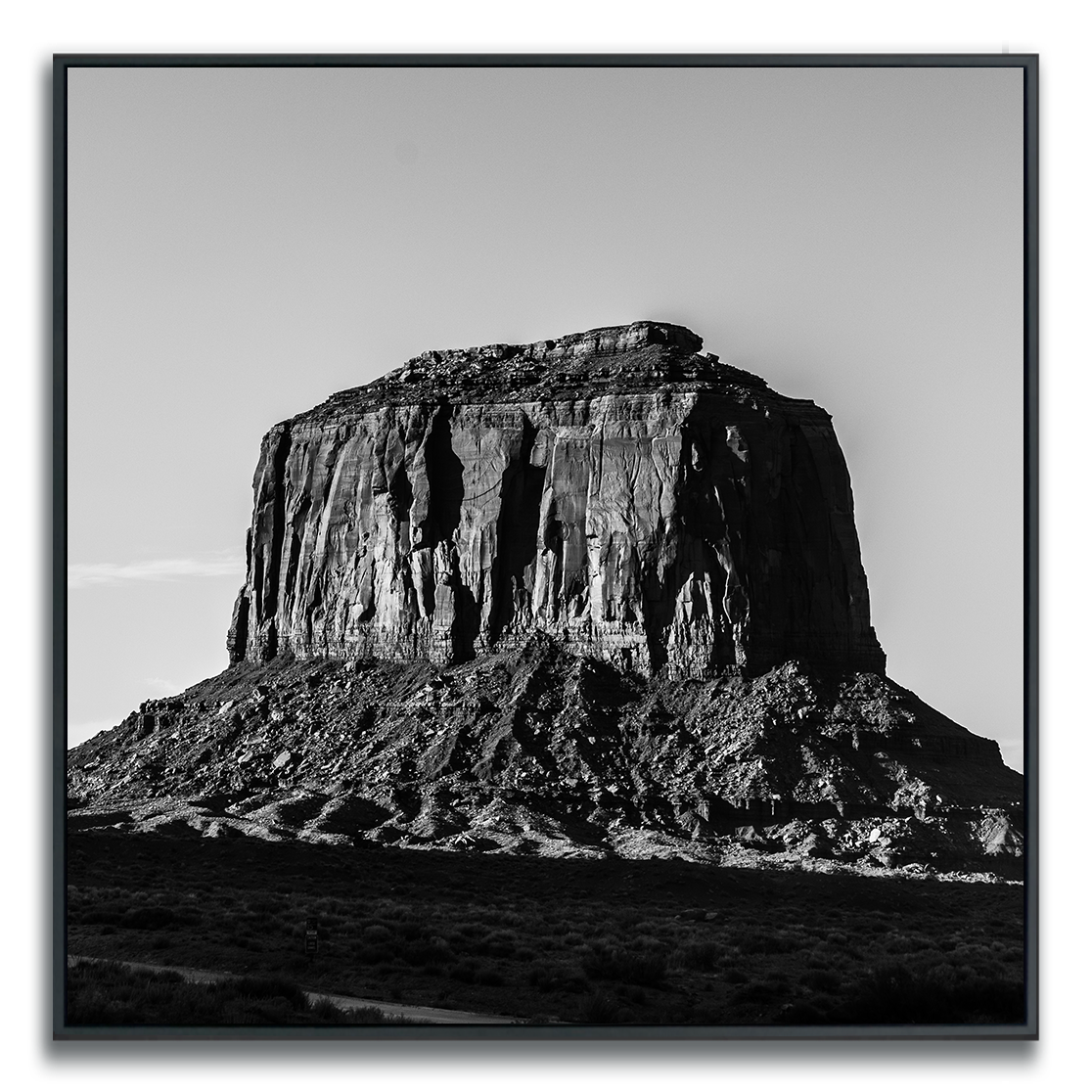 Black-and-white photograph of a prominent flat-topped rock formation rising from a barren landscape. The image highlights the butte’s steep, jagged cliffs and intricate surface textures, with dramatic light and shadow accentuating its monumental presence. Sparse vegetation and a clear sky enhance the sense of openness and isolation, evoking grandeur and timelessness.