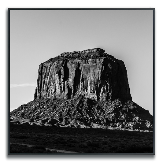 Black-and-white photograph of a prominent flat-topped rock formation rising from a barren landscape. The image highlights the butte’s steep, jagged cliffs and intricate surface textures, with dramatic light and shadow accentuating its monumental presence. Sparse vegetation and a clear sky enhance the sense of openness and isolation, evoking grandeur and timelessness.