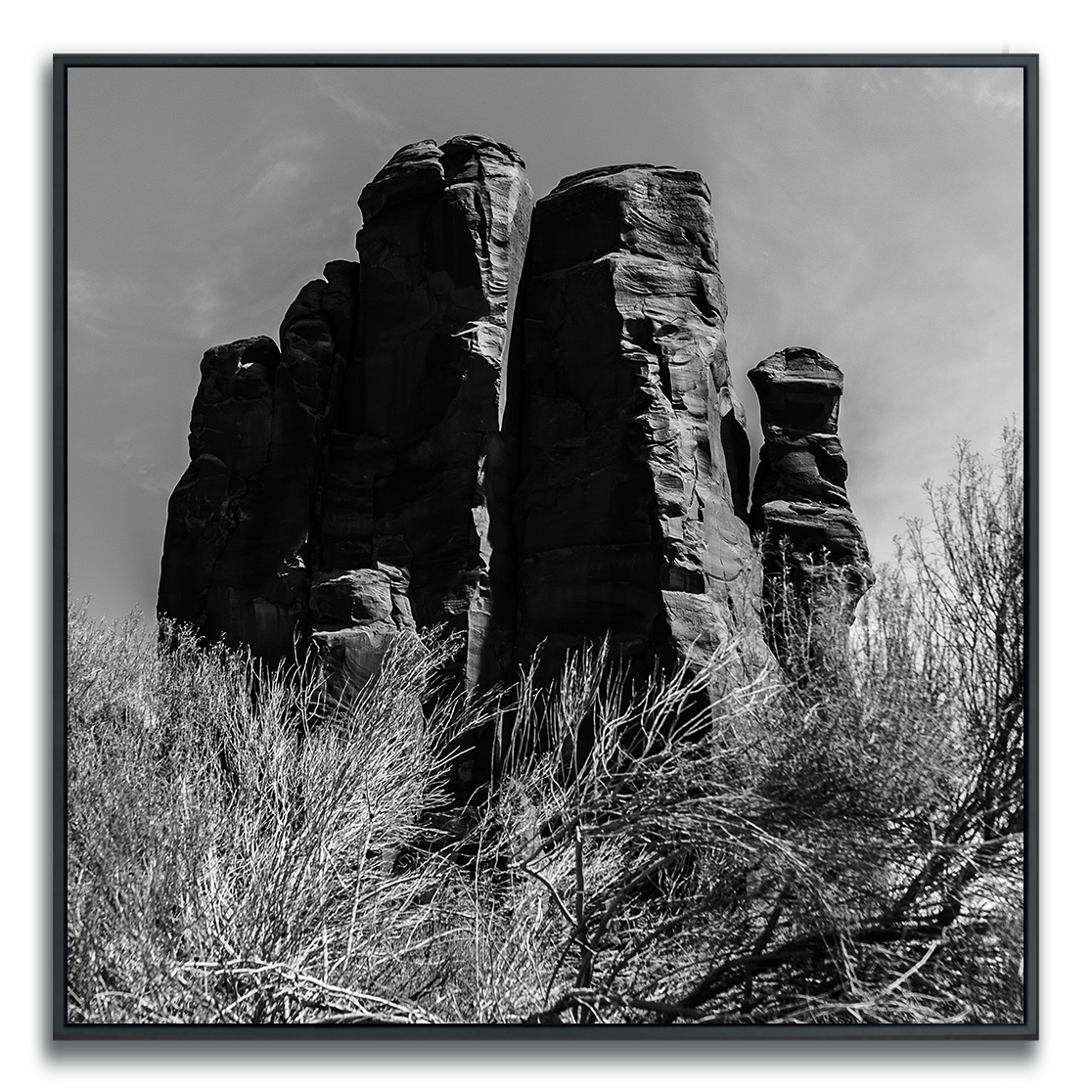 A black-and-white photograph featuring towering rock formations with rugged, vertical profiles. The dramatic silhouettes and textured surfaces are accentuated by strong light and shadow, while sparse brush at the base adds a touch of softness. The vast sky and open landscape evoke a sense of grandeur, isolation, and timeless natural beauty.