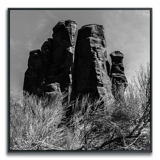 A black-and-white photograph featuring towering rock formations with rugged, vertical profiles. The dramatic silhouettes and textured surfaces are accentuated by strong light and shadow, while sparse brush at the base adds a touch of softness. The vast sky and open landscape evoke a sense of grandeur, isolation, and timeless natural beauty.