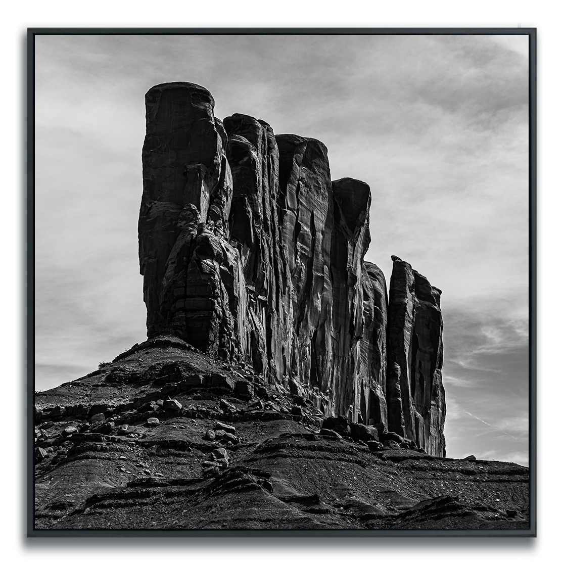 A striking black-and-white photograph of a dramatic rock formation with towering, vertical cliffs rising against a softly clouded sky. The rugged textures and deep grooves in the stone are highlighted by the interplay of light and shadow, creating a sense of grandeur, isolation, and timelessness in the natural landscape.