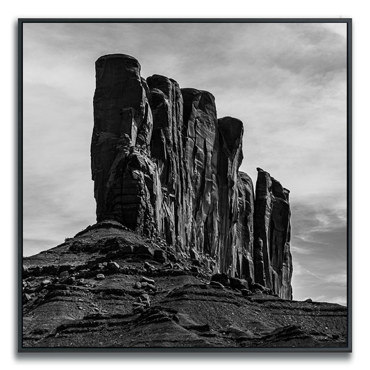 A striking black-and-white photograph of a dramatic rock formation with towering, vertical cliffs rising against a softly clouded sky. The rugged textures and deep grooves in the stone are highlighted by the interplay of light and shadow, creating a sense of grandeur, isolation, and timelessness in the natural landscape.