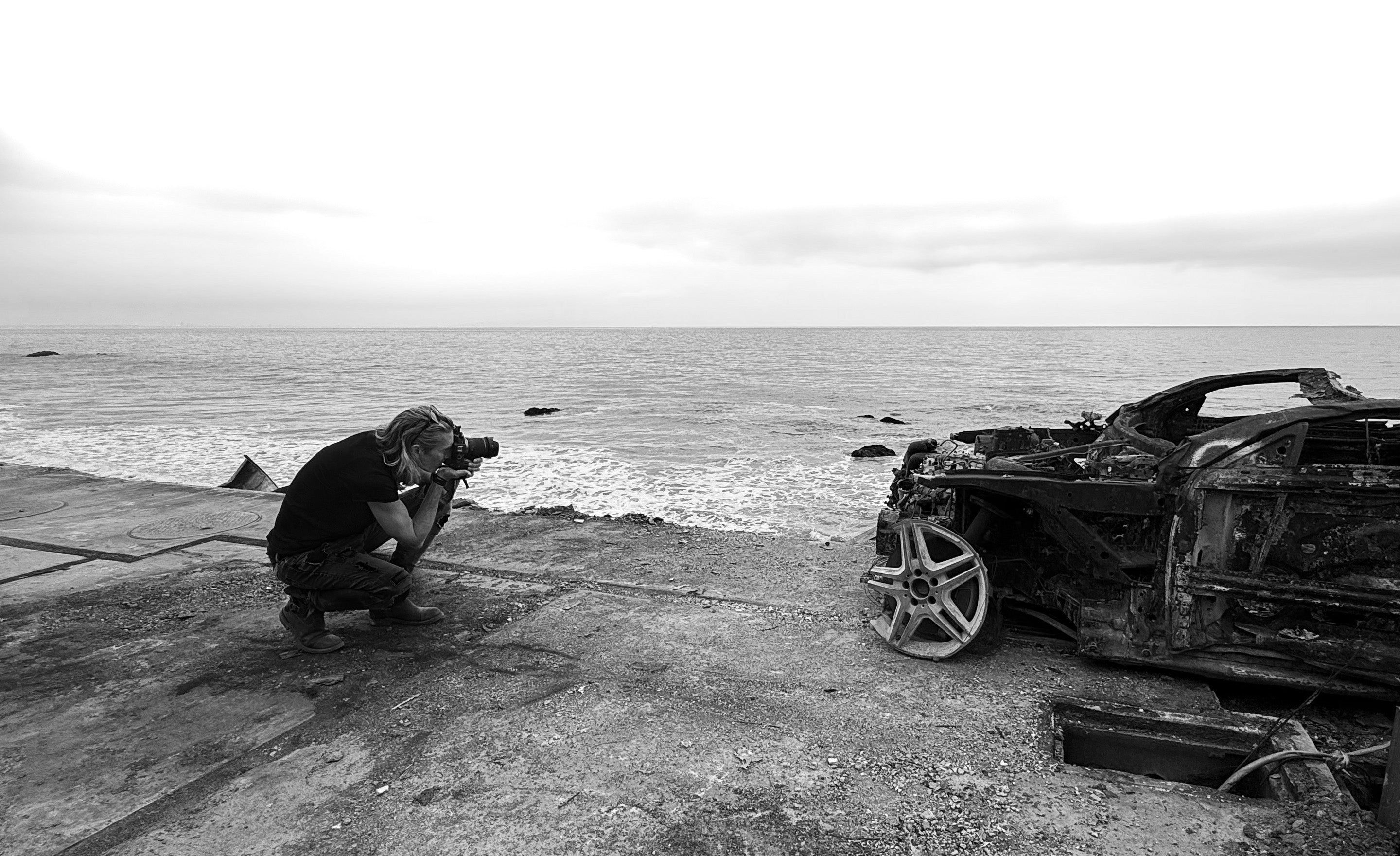 Los Angeles Fine Art Photographer Mark Maryanovich promotional Image kneeling by ocean photographing burnt car holding camera black and white from side perspective