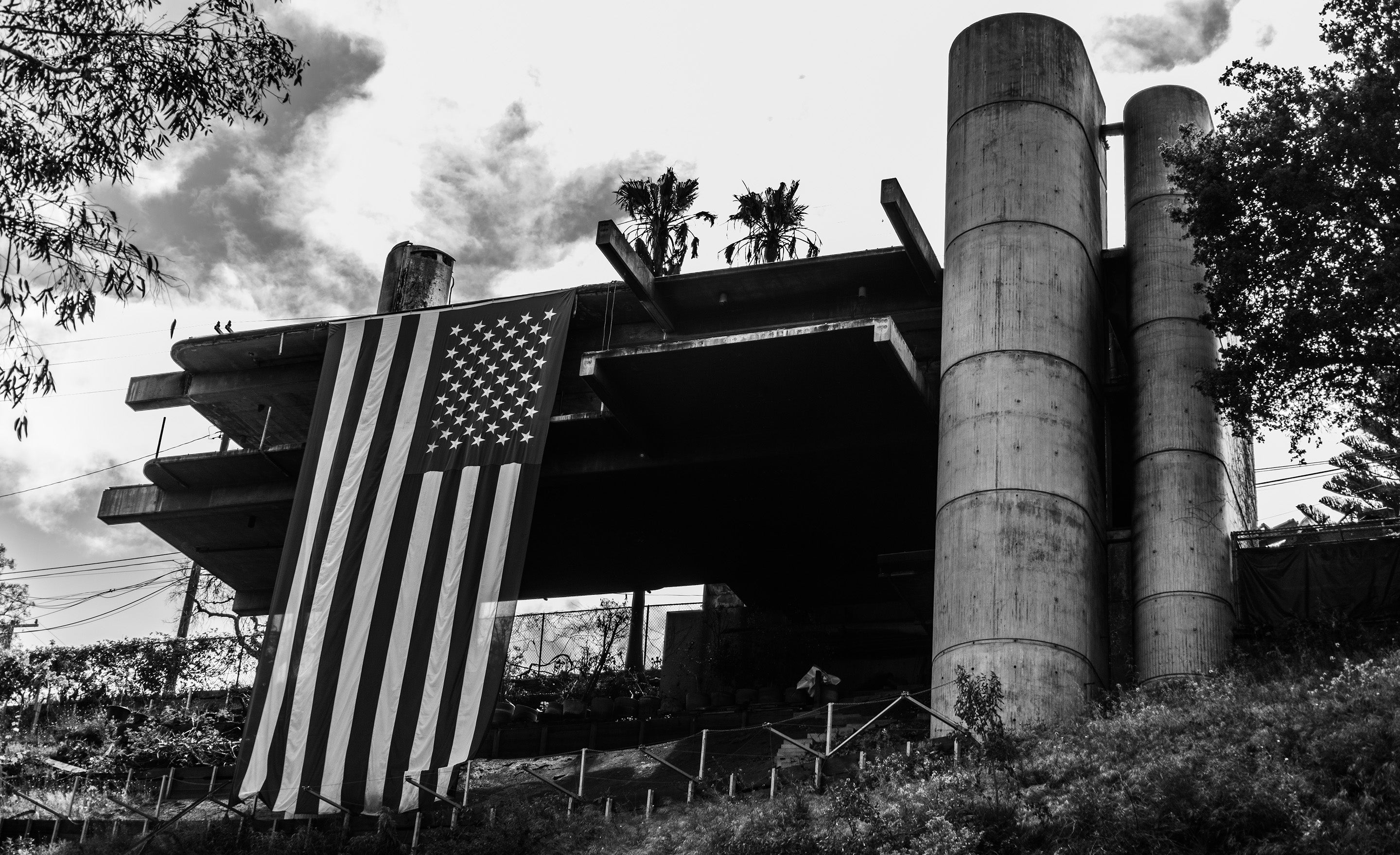 Black and white fine art photograph burnt house with American Flag hanging of deck with clouds behind