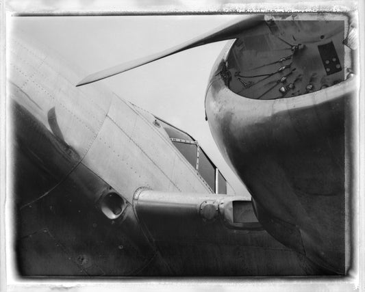 Black and white close-up photograph of an airplane engine and propeller, presented with a distressed vintage border effect.