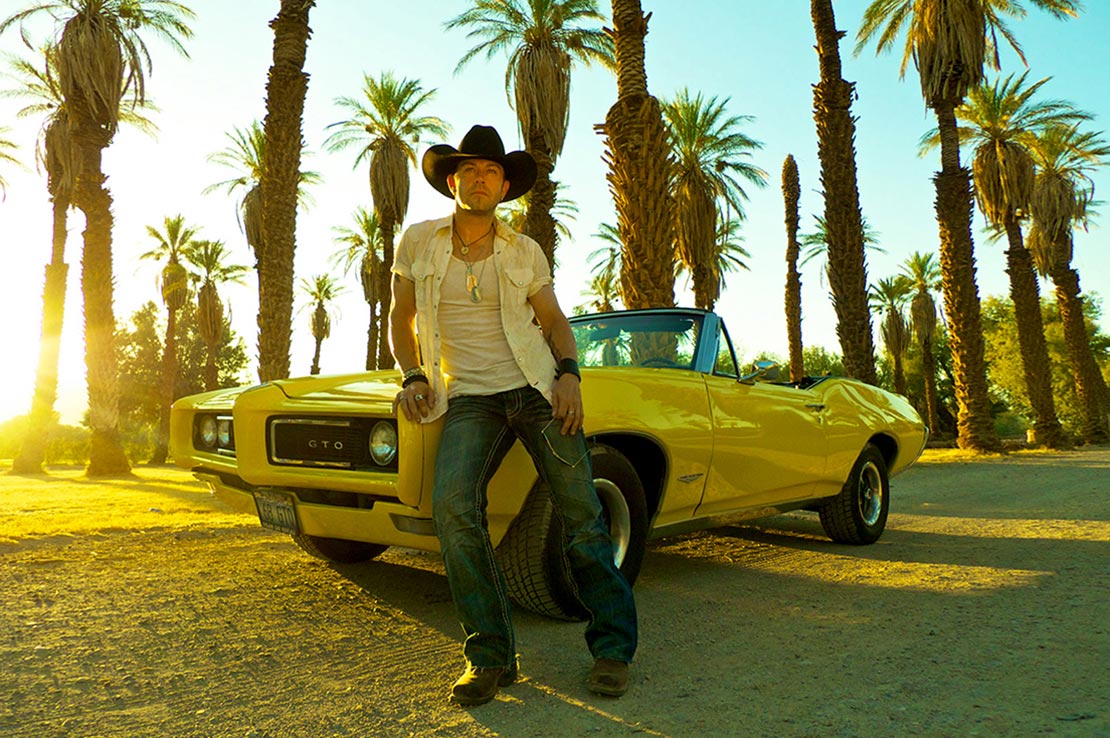 Male musician portrait Aaron Pritchett wearing jeans white shirt black cowboy hat leaning against yellow convertible front grill palm tress blue sky background