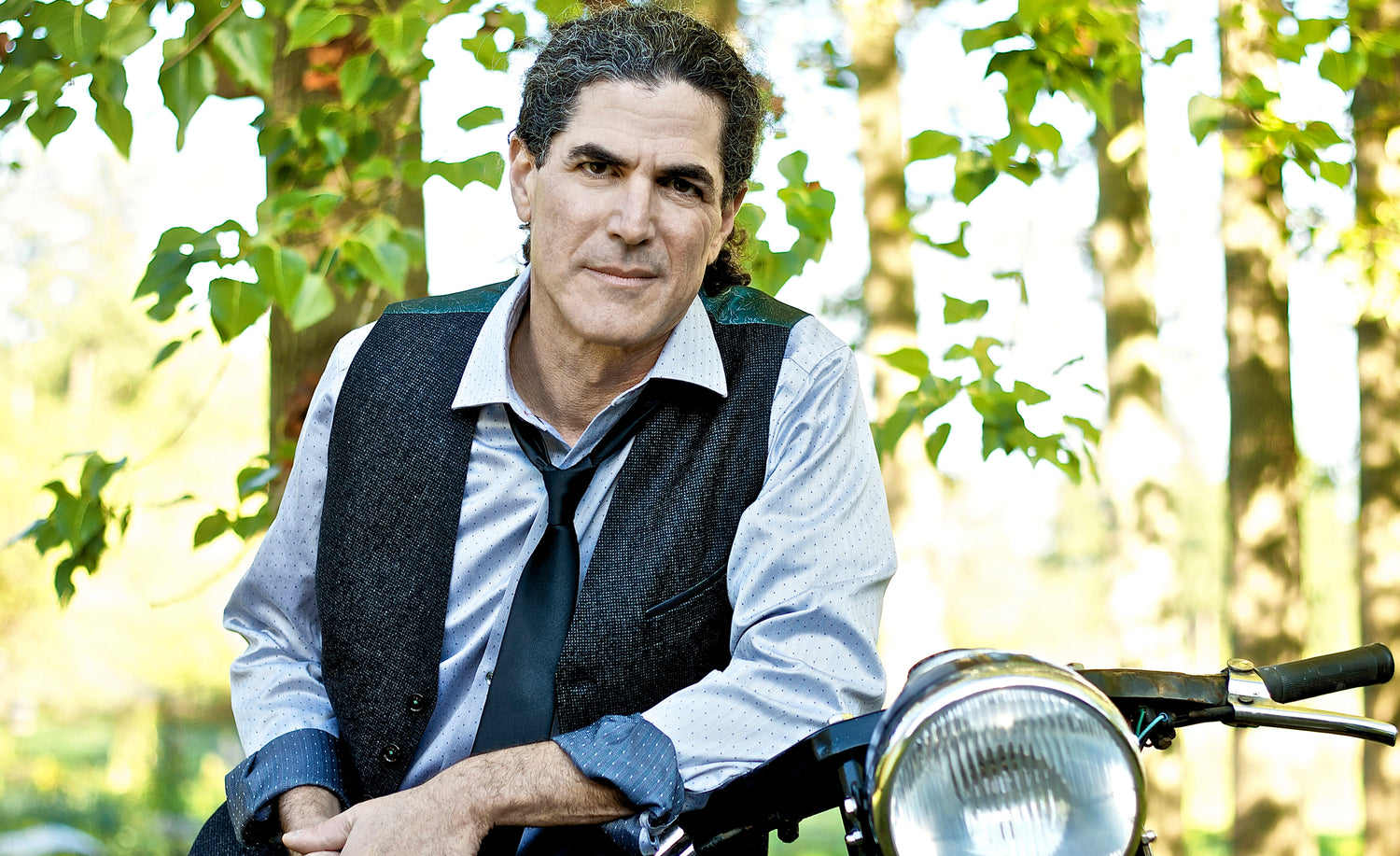 Male portrait  Andrew Riseman wearing vest and tie leaning against motorcycle handlebar with trees behind