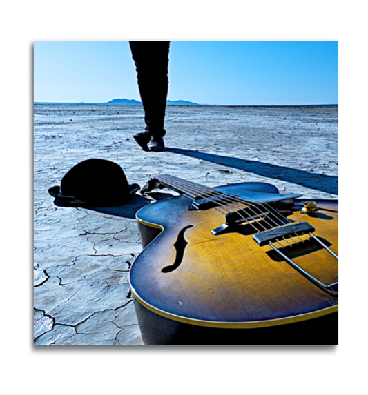 Fine art square color photographic print on metal Gibson acoustic vintage guitar lying on desert dried lakebed in foreground with hat beside it legs with feet crossed in background