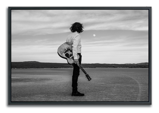 Black and white Fine Art Photography man standing in desert holding Gibson acoustic guitar back to camera staring at moon above the horizon