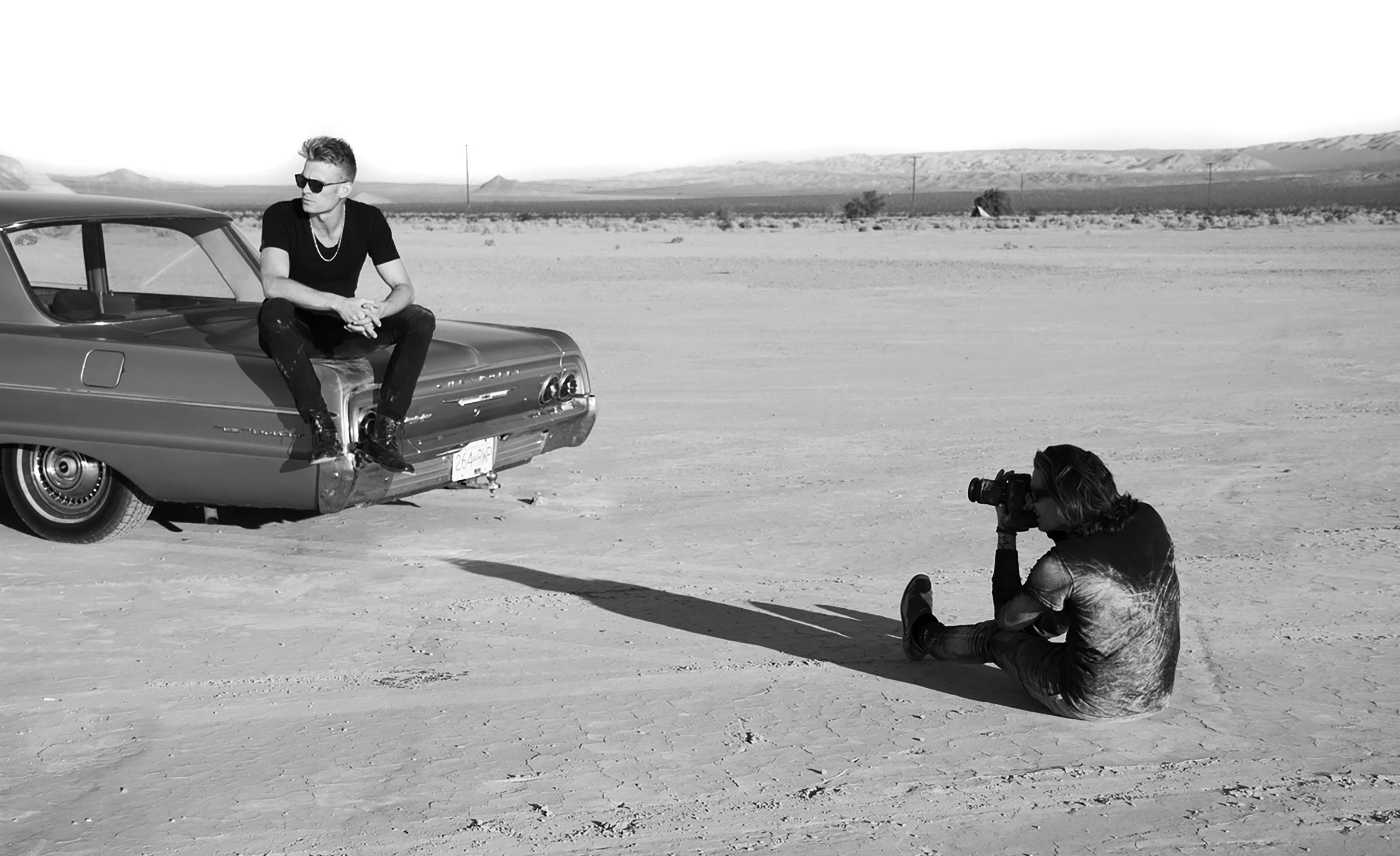 Black and white image man wearing black sitting on car trunk in desert having his photo taking