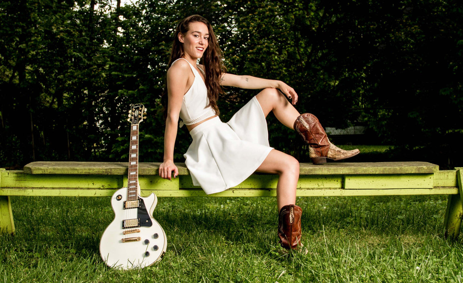 Country Music portrait Nashville Leanne Pearson sitting on bench in white dress with cowboy boots white electric guitar leaning beside her  trees behind her grass below