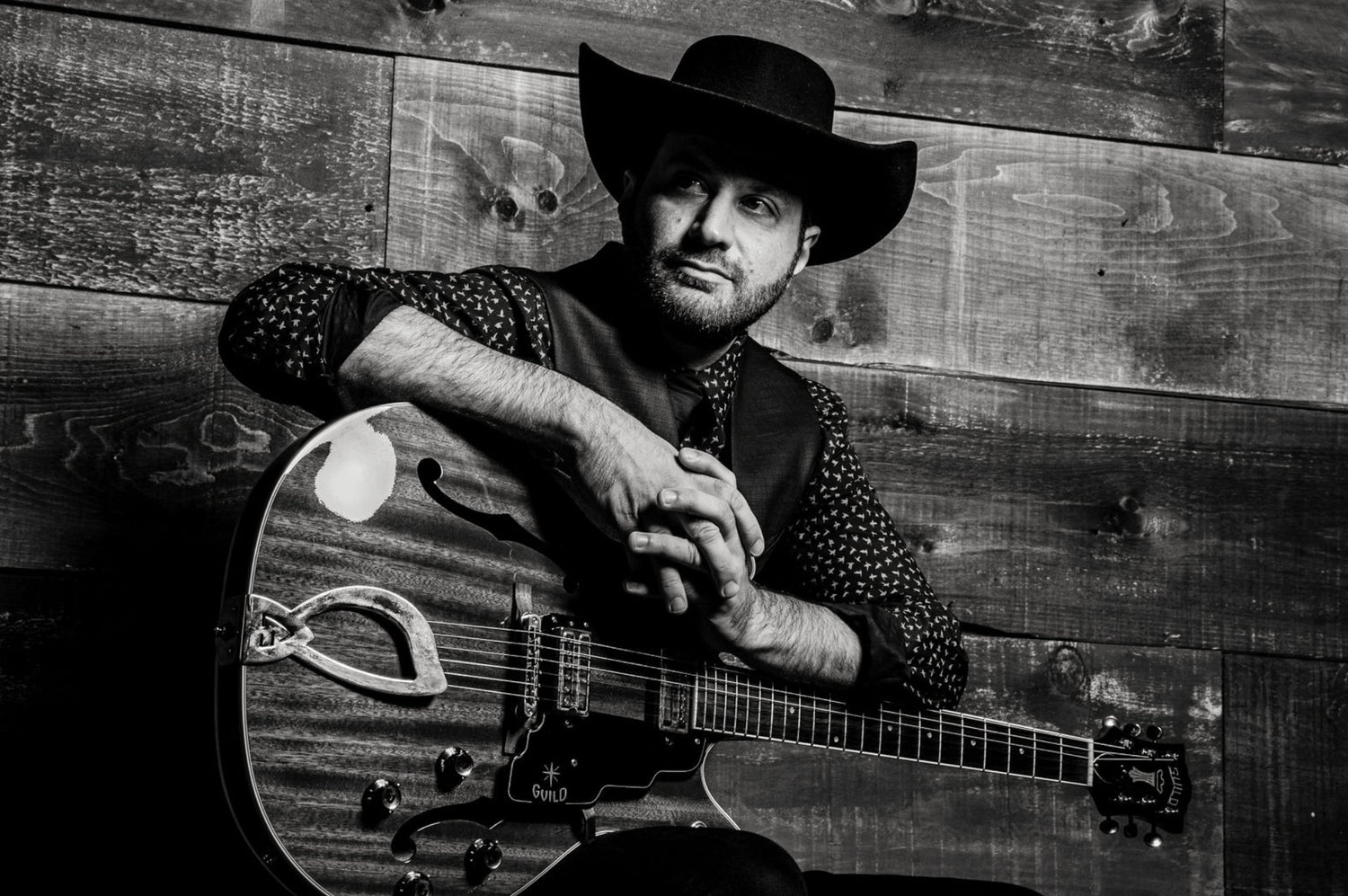 Portrait Country Music Artist Doc Reinhardt New York sitting against wood wall with guitar in his lap and black cowboy hat