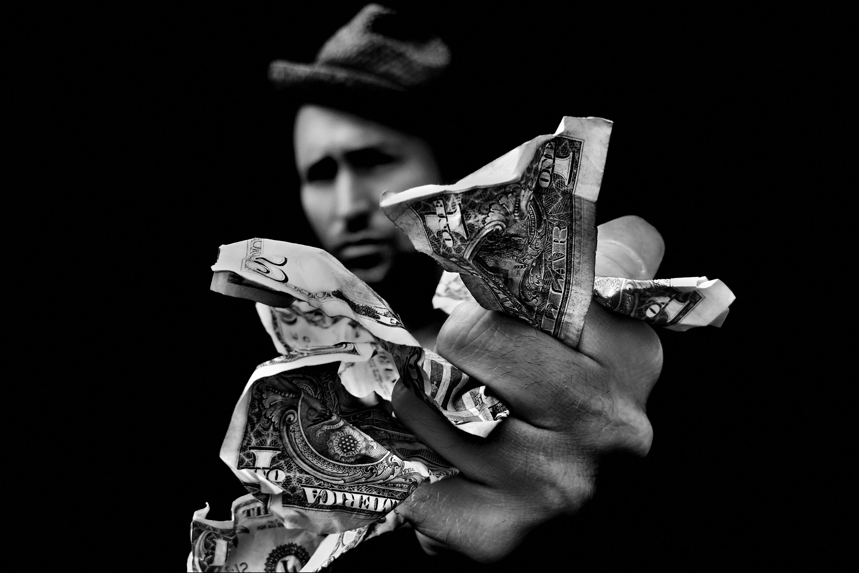 Black‑and‑white photograph of a hand gripping crumpled dollar bills in front of a blurred figure.