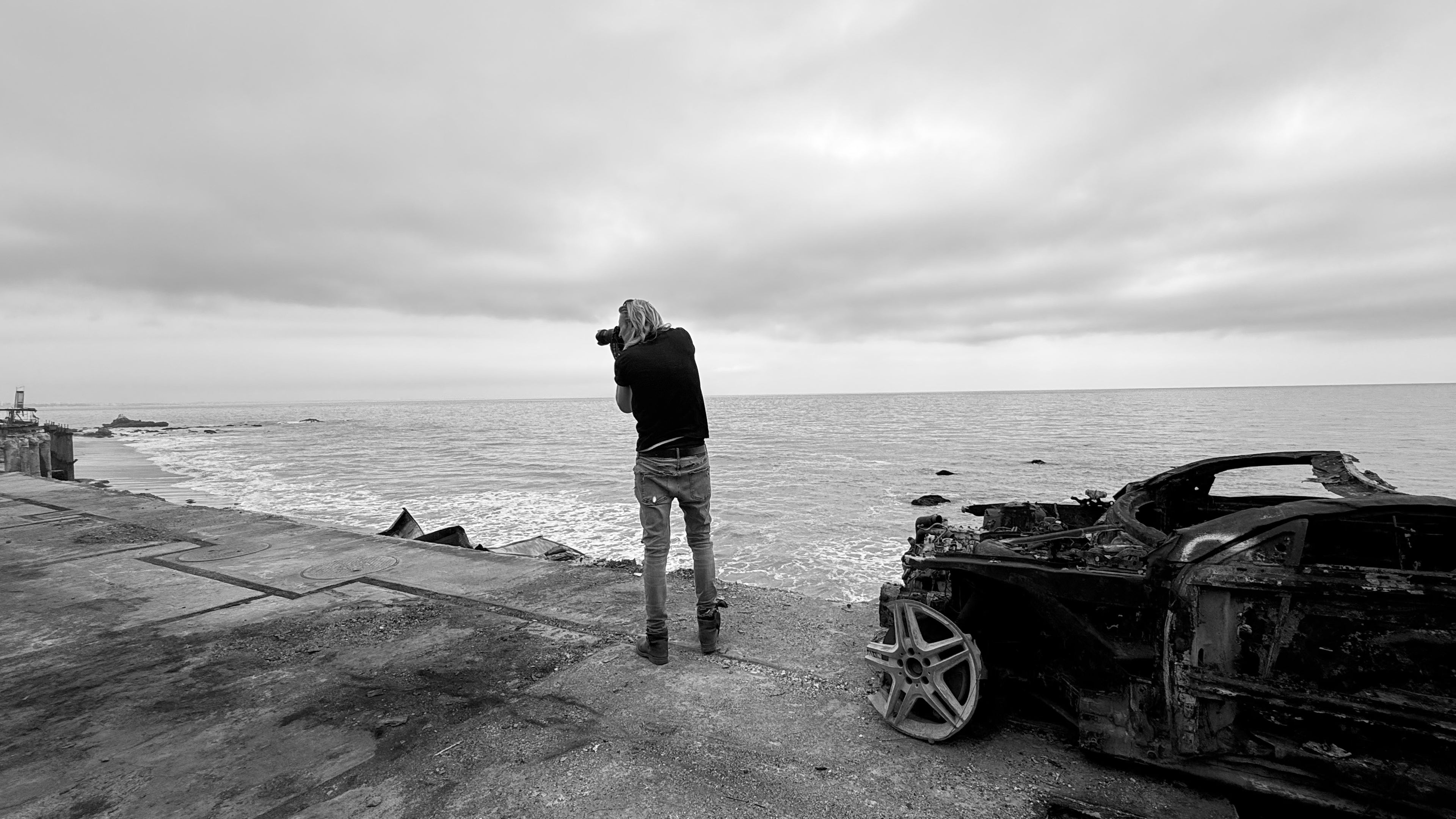 Fine Art Photographer Mark Maryanovich taking a photo by a burned out car by the sea on a cloudy day in black and white.
