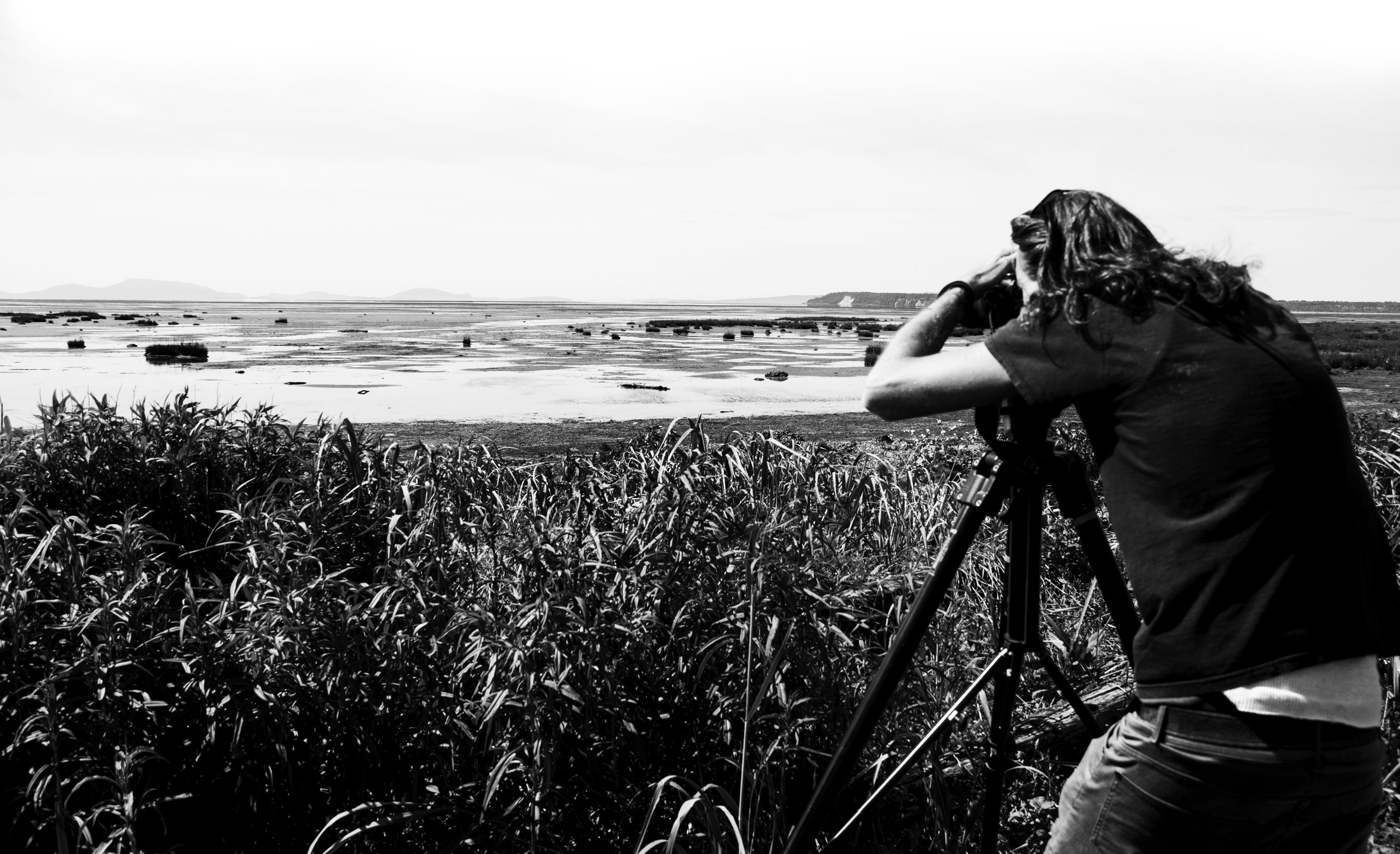 Fine art photographer Mark Maryanovich behind the scenes black and white photo looking through back of camera on tripod behind long grasses overlooking a dyke at low tide