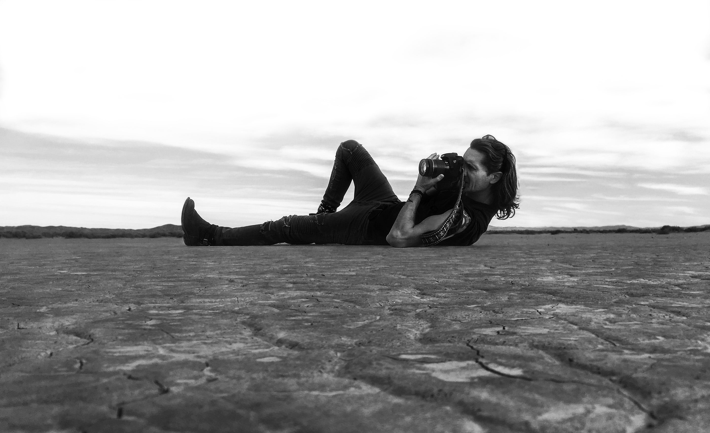 Black and white photo Mark Maryanovich lying n dry lake bed holding camera