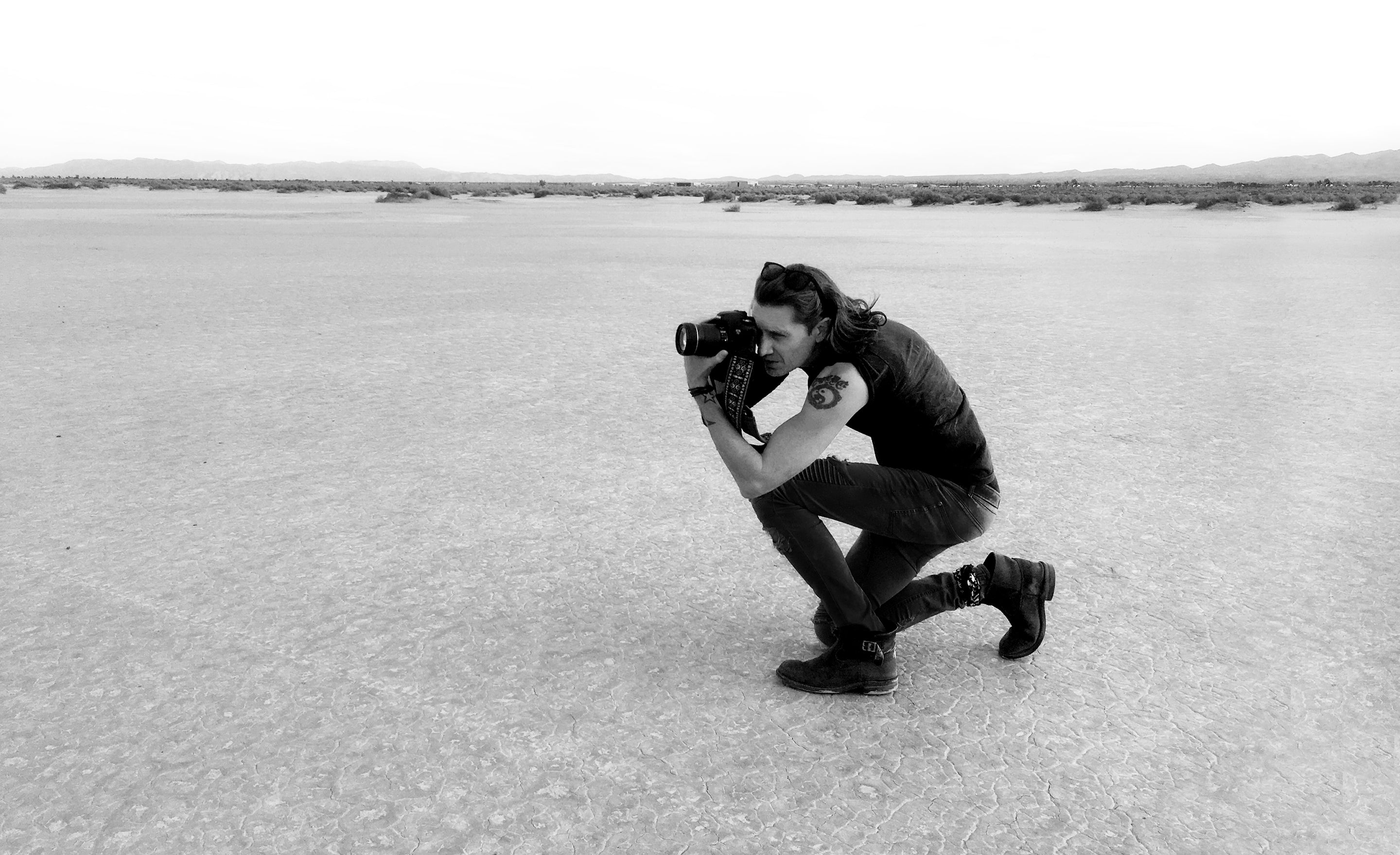 Fine art photographer Mark Maryanovich kneeling on dried lake bed floor looking through back of camera in black and white