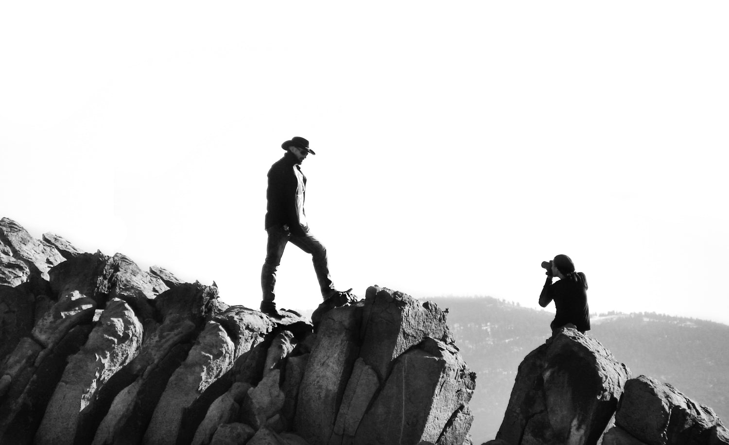 Black and white photo of Mark Maryanovich photographing by Todd Richard standing on rocky cliff wearing cowboy hat