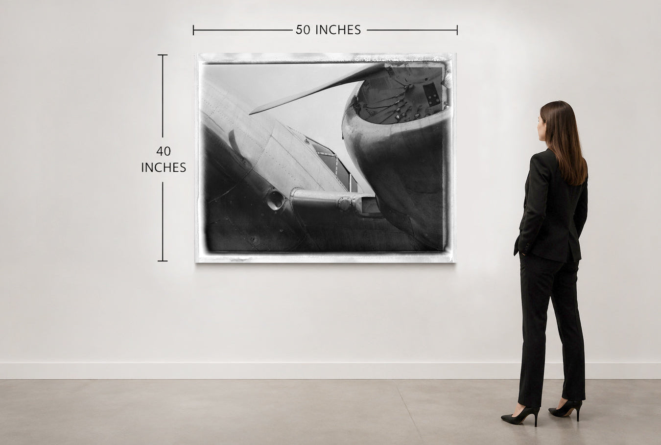 A woman in a black suit stands in a gallery room looking at a framed black-and-white airplane photograph mounted on a white wall. Measurement lines and text indicate the print is 50 inches wide by 40 inches tall.