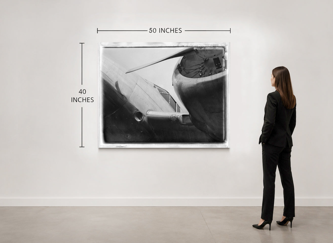 A woman in a black suit stands in a gallery room looking at a framed black-and-white airplane photograph mounted on a white wall. Measurement lines and text indicate the print is 50 inches wide by 40 inches tall.