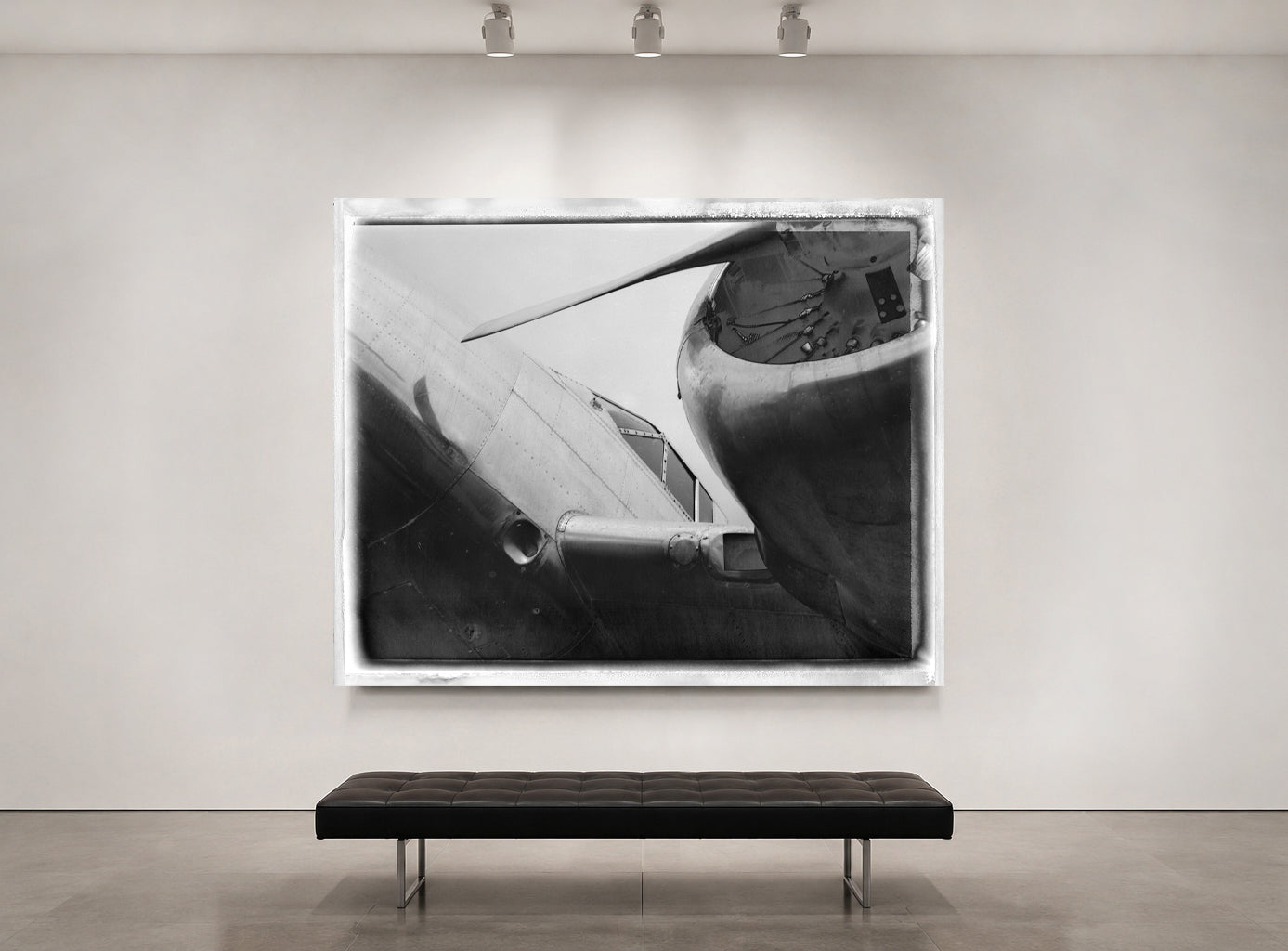 A large black and white framed art print depicting a close-up of an airplane engine and wing, displayed on a light gray gallery wall above a dark tufted bench.