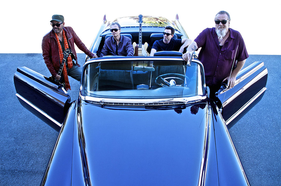 Music group portrait Harpdog Brown And The Travelin Blues Show two members standing on either side of classic car with doors open two other members in back seat all wearing sunglasses