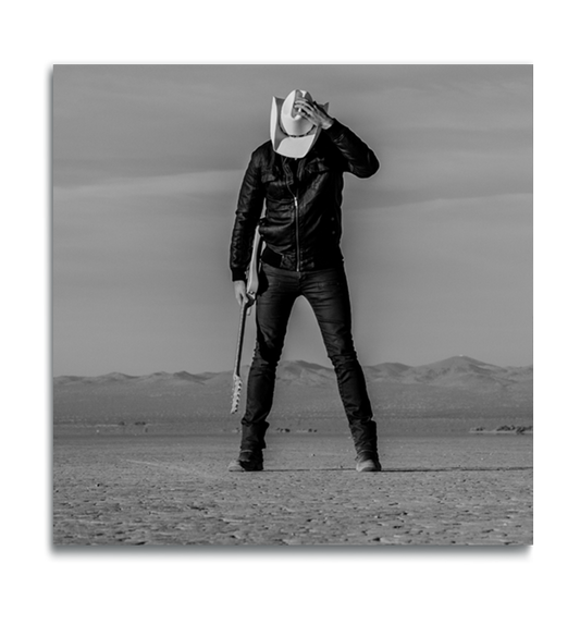 Black and white square metal print man standing in desert holding guitar head down holding onto his white cowboy hat covering his face