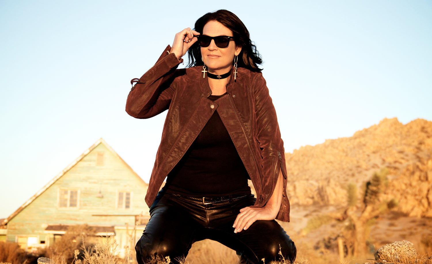 Female musician portrait Leslie Cours Mather on knees in desert on hand to her sunglasses mountain and building behind her