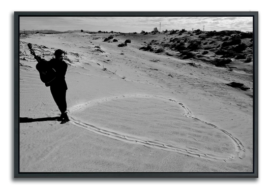 Black and white fine art photography man standing with guitar at the point of a heart drawn in sand