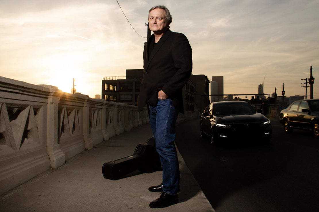 Musician portrait Mangos Daddy standing on bridge at sunset traffic beside him one hand in pocket and black hard guitar case on sidewalk behind him