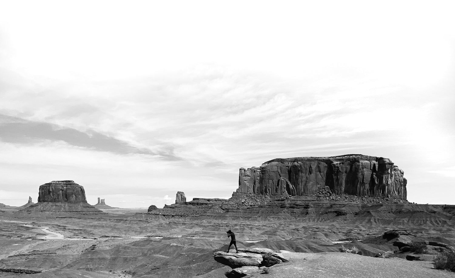 Panoramic shot in black and white, fine art photographer Mark Maryanovich standing on precipice at John Ford Point, dwarfed by the desert buttes of Monument Valley, aiming his camera at the horizon.