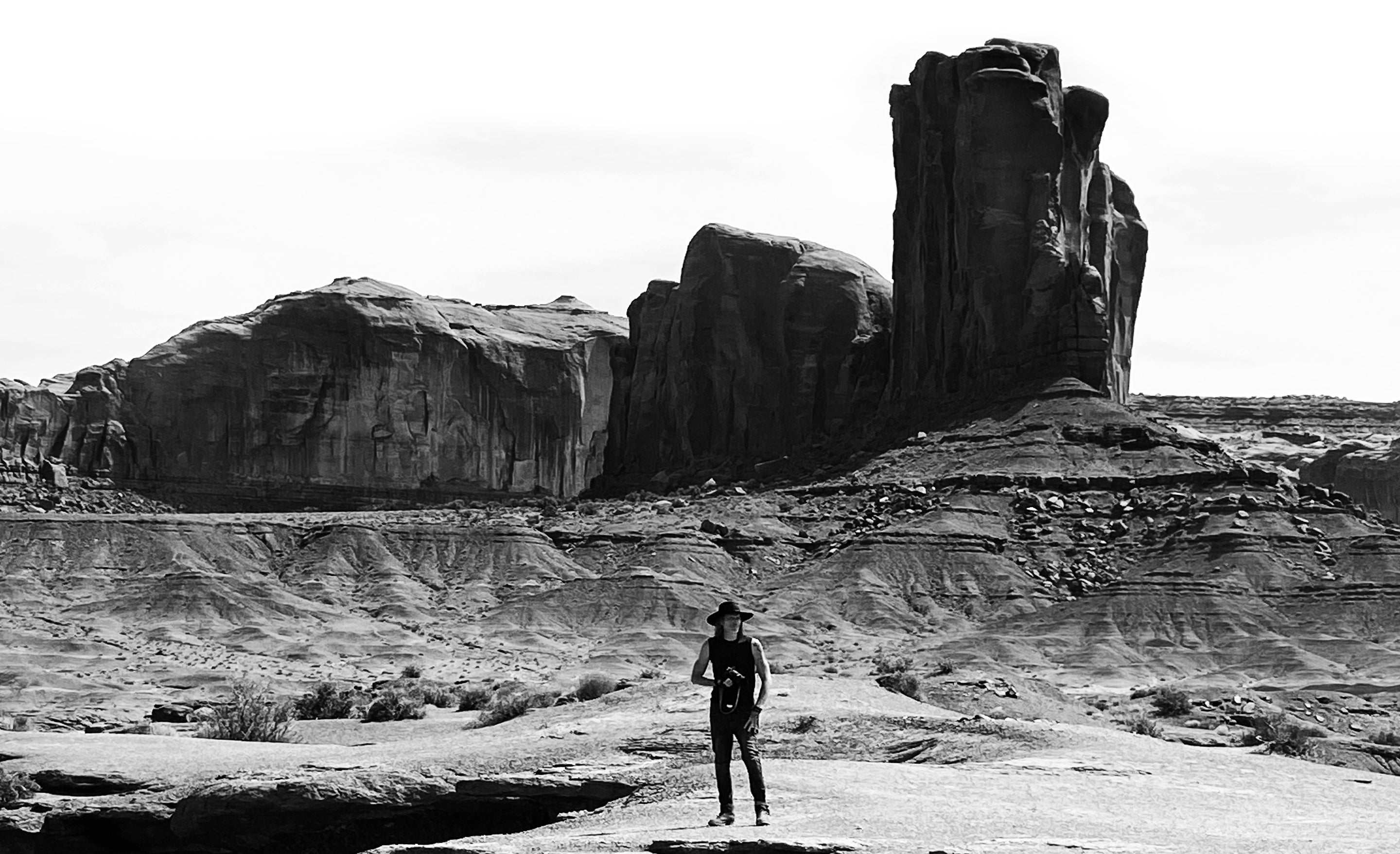 Black and white photo Mark Maryanovich standing beneath cliff in Monument Valley holding camera