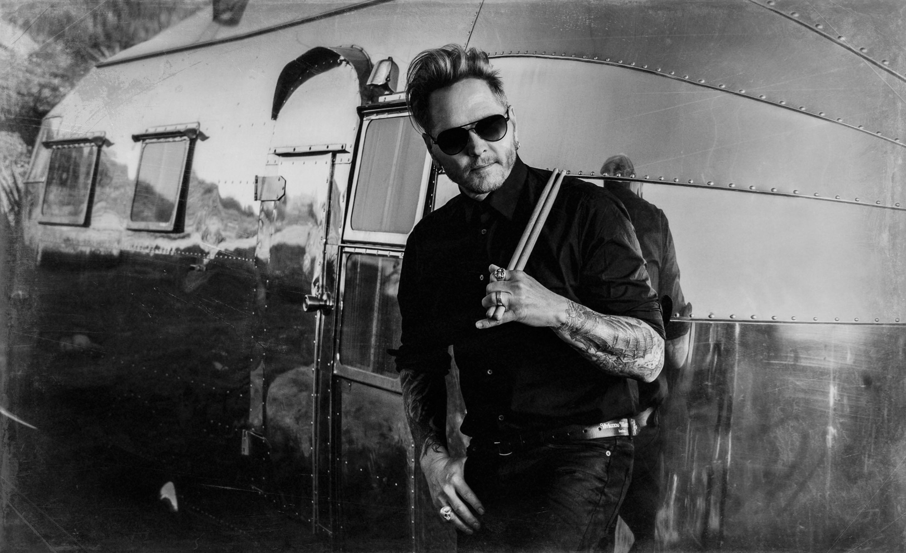 Matt Sorum musician black and white portrait standing against silver airstream trailer holding drumsticks that are leaned on his shoulder other hand hanging off his pants pocket