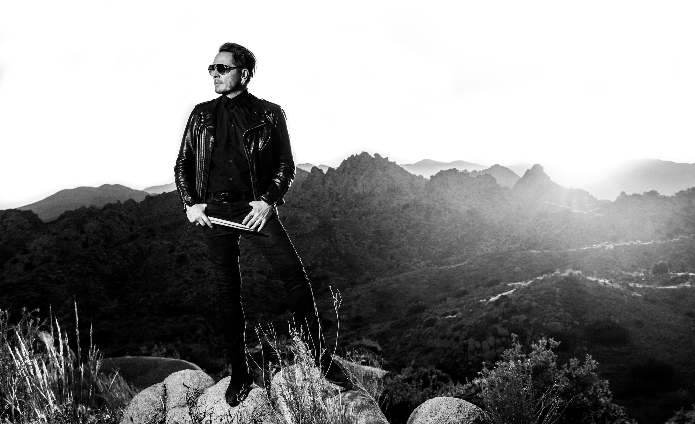 Matt Sorum black and white portrait on hill top in desert valley wearing sunglasses while holding drumsticks thumbs in his denim pockets