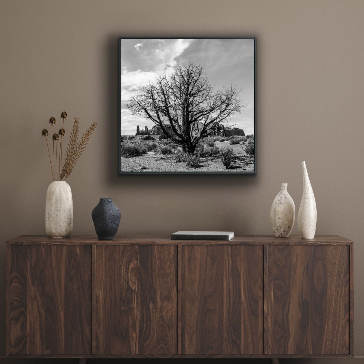 Black and white photograph of a lone dried tree in foreground, Monument Valley in distance framed print above a wooden sideboard with decorative vases.