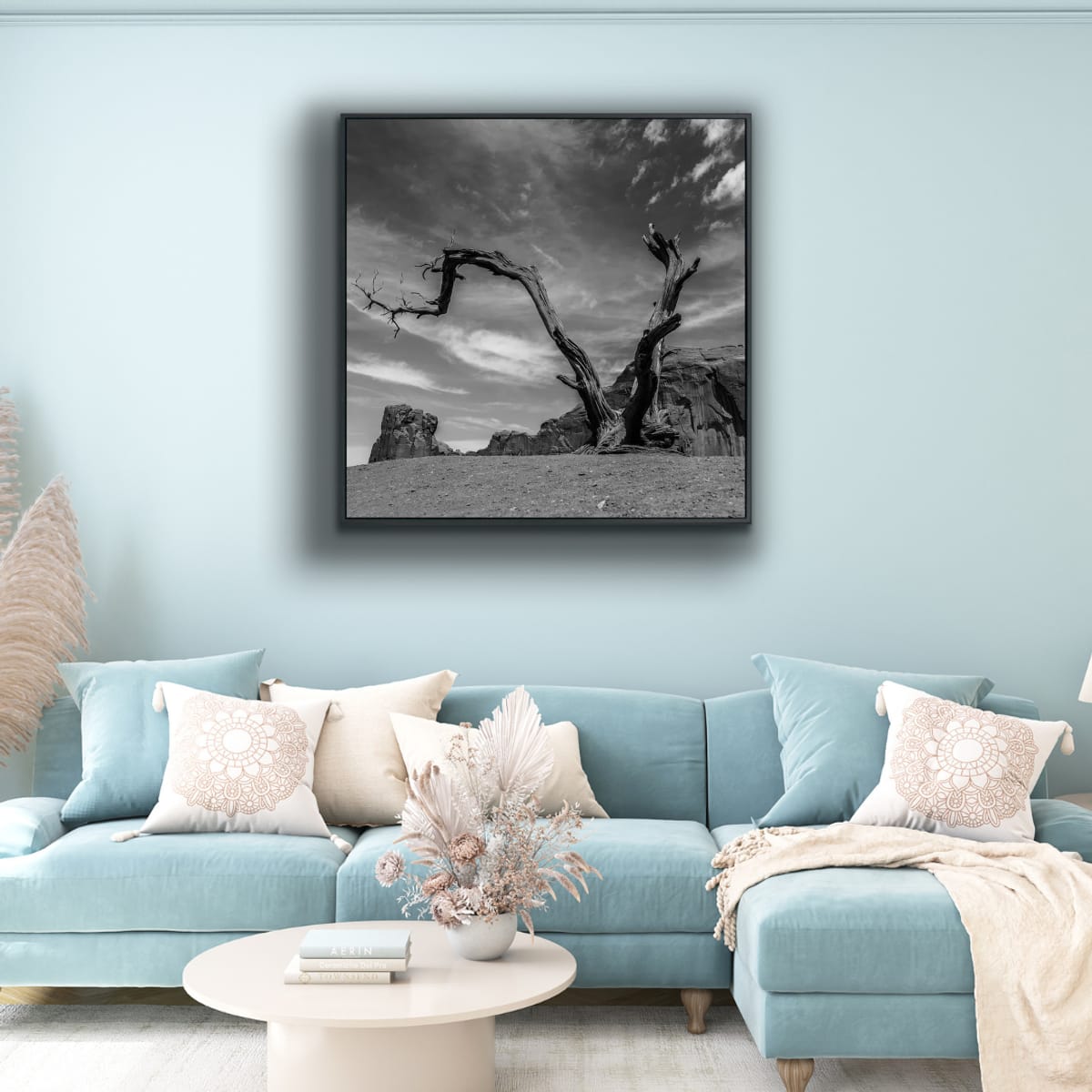 Light blue sofa with decorative pillows and a coffee table in a living room, featuring a dead tree with twisted branches against a dramatic sky and rocky landscape in Monument Valley