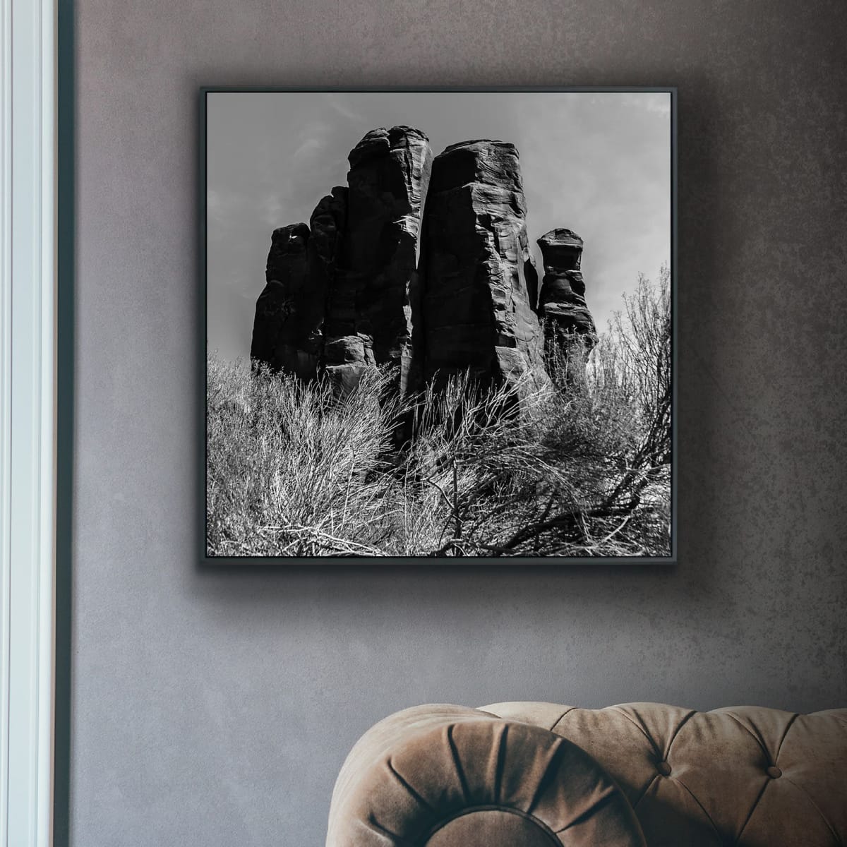 Framed black and white photograph of Black and white photograph of rock formations with desert vegetation on a textured gray wall above a brown sofa.