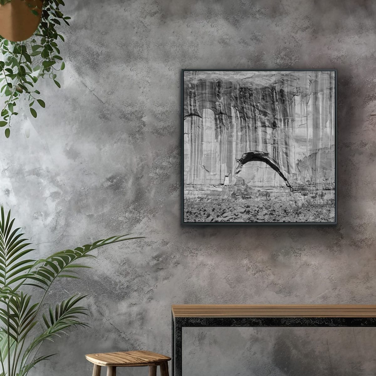 Black and white large square photograph of a Monument Valley rock formation with arc on a textured wall with plants and a wooden narrow long table and stool.
