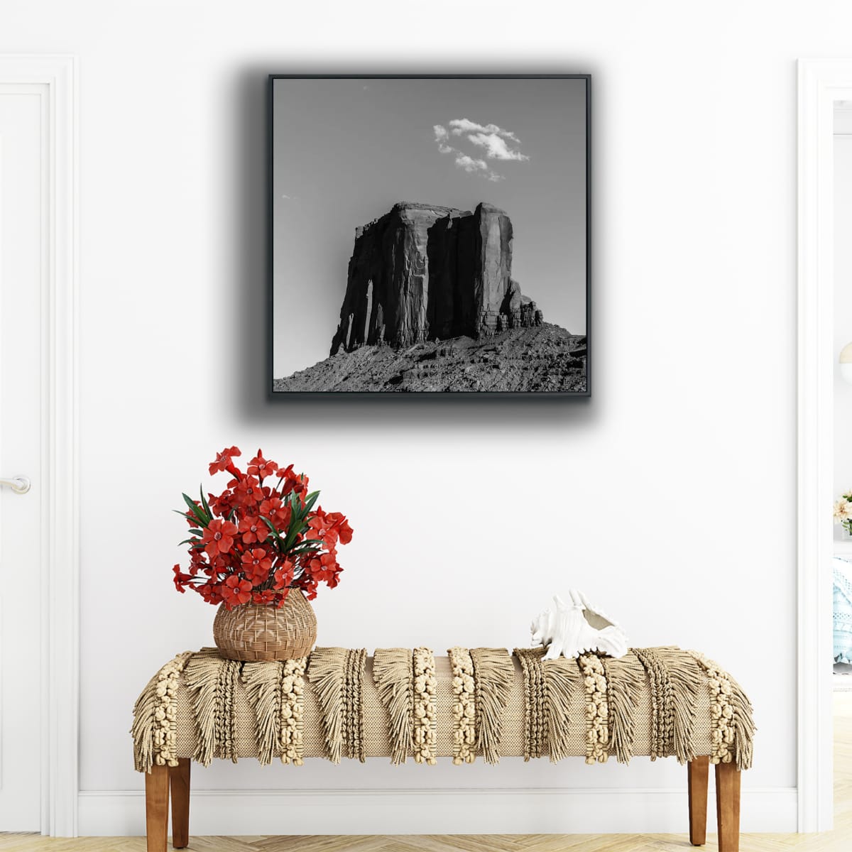 Framed black and white 48x48 Monument Valley artwork in black and white of a solitary butte beneath a small cloud formation, hanging on a white wall above a long bench with potted plant with red flowers placed on it in a home foyer.