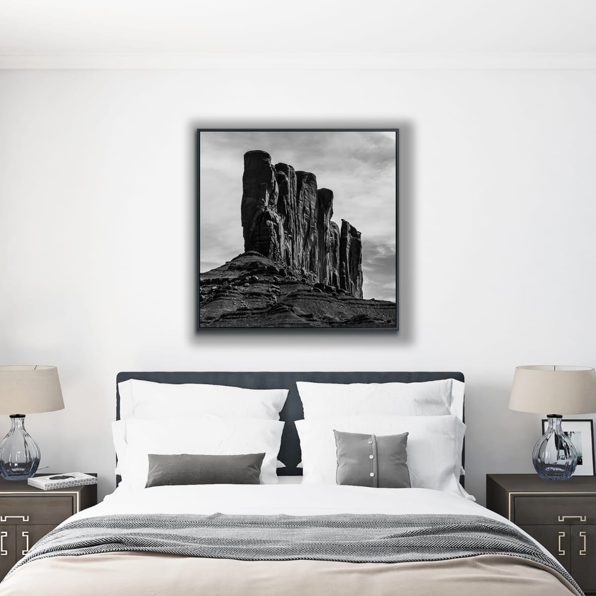Framed black and white photograph of a rocky landscape with tall rock formations against a cloudy sky above a bed in a modernly styled bedroom.
