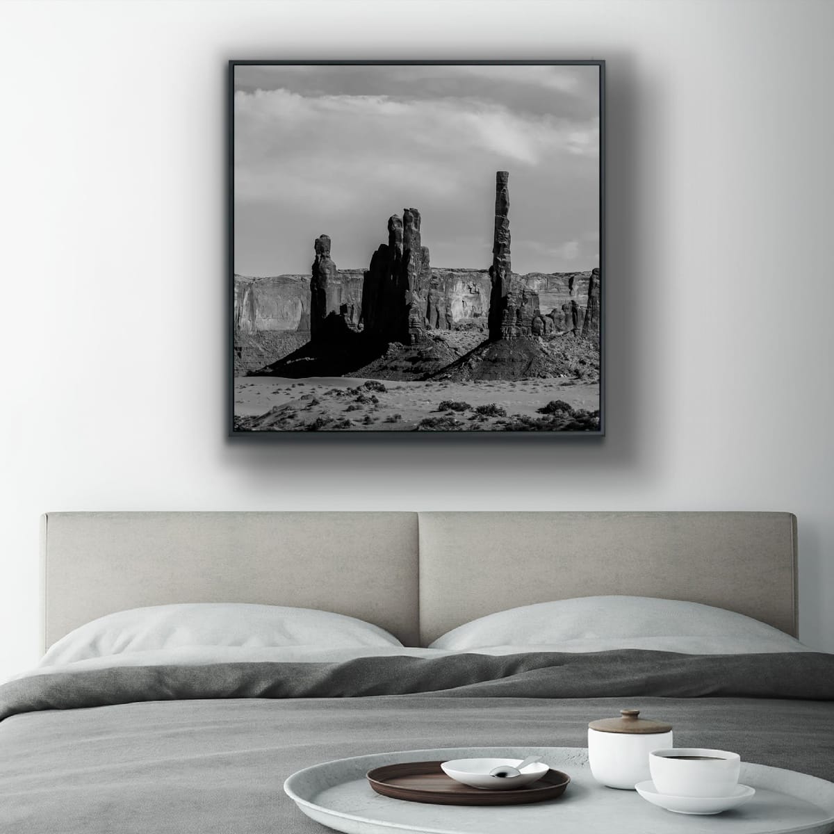 Framed black and white square print of Monument Valley desert butte spires above a hotel room bed with a minimalistic decor and breakfast tray on the bed in the foreground.