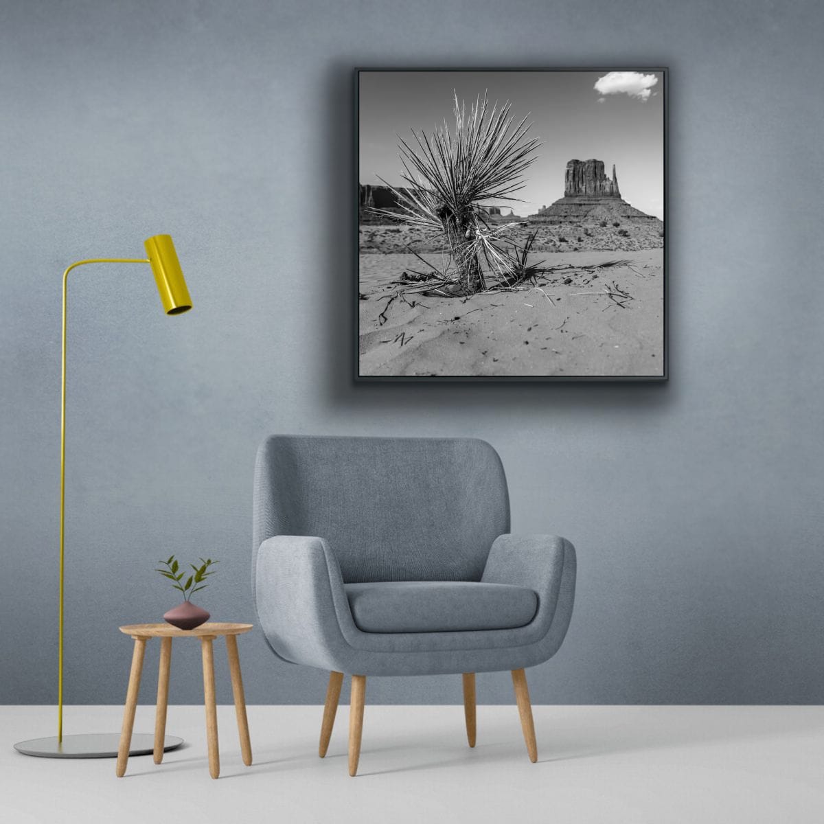 Modern living room with a gray armchair, yellow floor lamp, and framed black and white landscape on the wall featuring a square black and white photograph of a desert landscape with a cactus and iconic Monument Valley butte in the background.