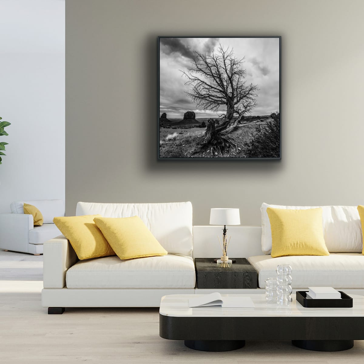 Modern living room with white sofa, yellow cushions, and a framed black and white photograph on the wall, featuring a lone curved tree in foreground, iconic Monument Valley buttes in background.