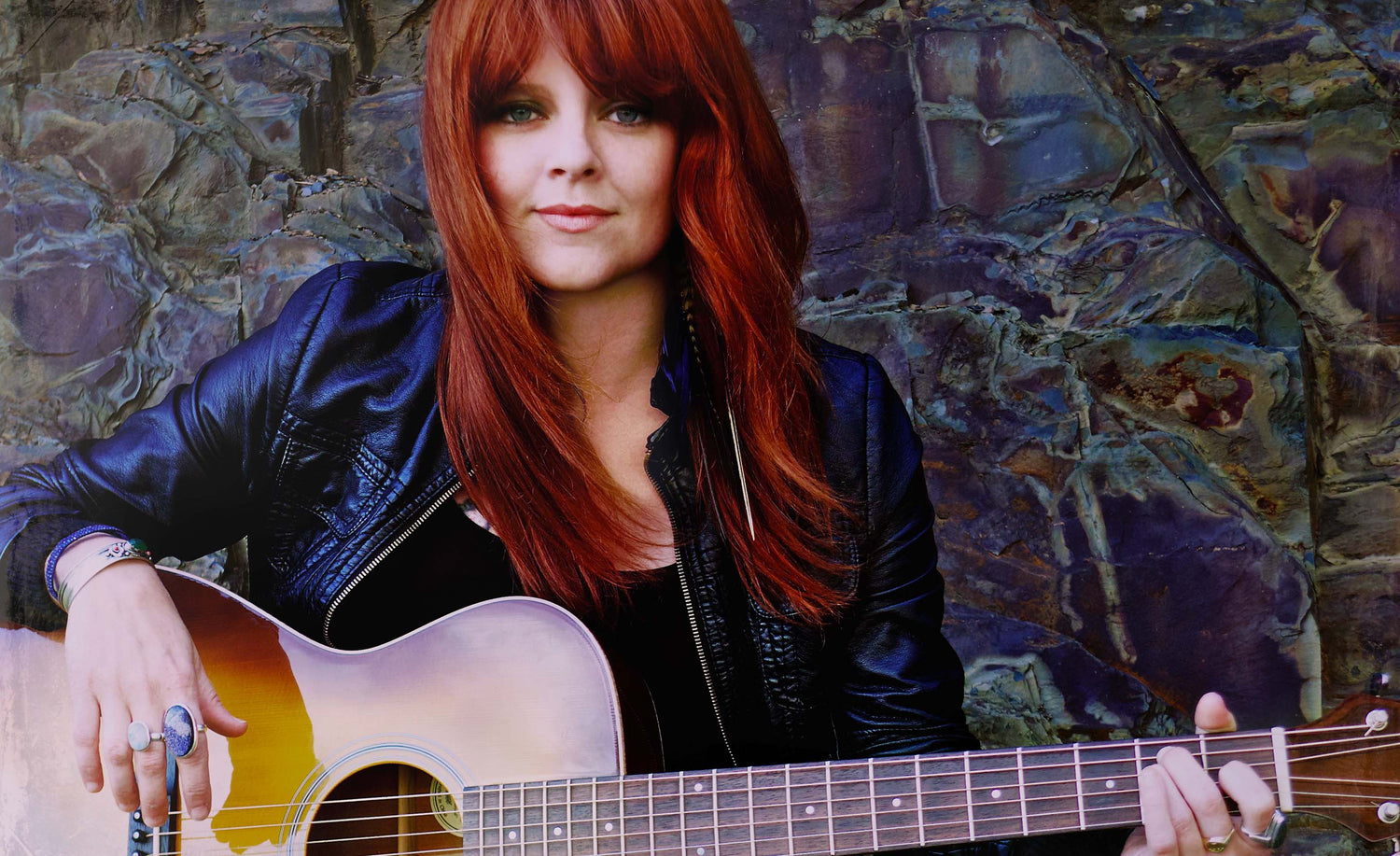 Music artist portrait Carmen Townsend sitting with acoustic guitar in lap against green stone wall