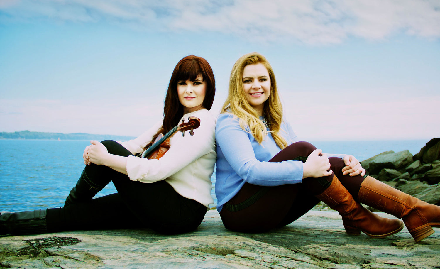 Music Duo photo Cassie and Maggie sitting on rocks knees bent ocean behind them