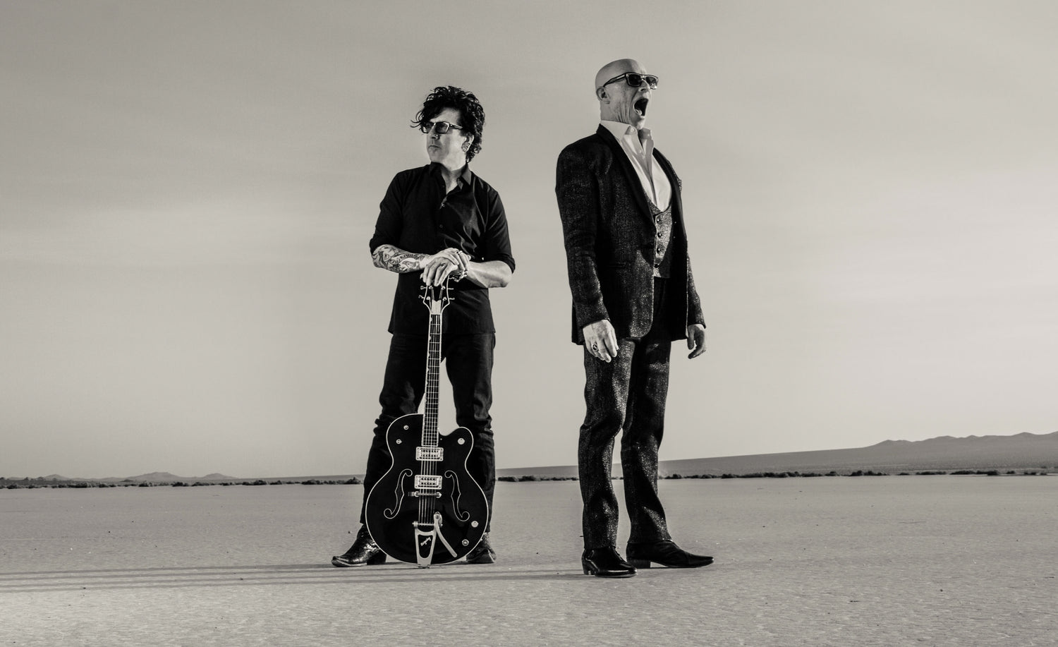 Black and white band portrait horse duo standing in desert one member has hands resting on neck of huitar standing upright between his leg singer drummer looking away with mouth open El Mirage