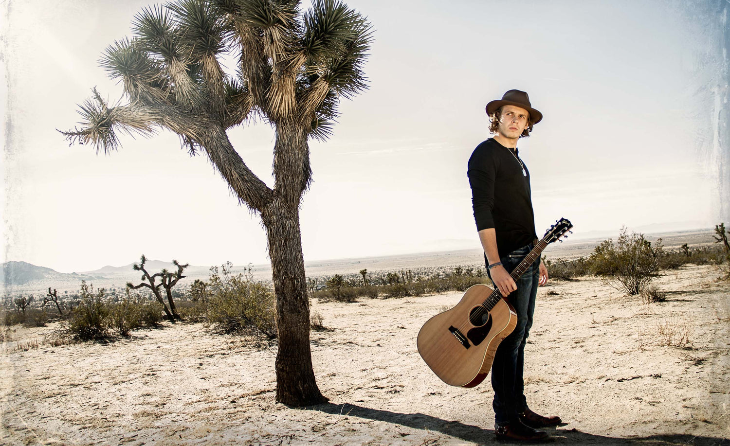 Musician photo Cole Bradley standing in desert by Joshua Tree holding acoustic guitar