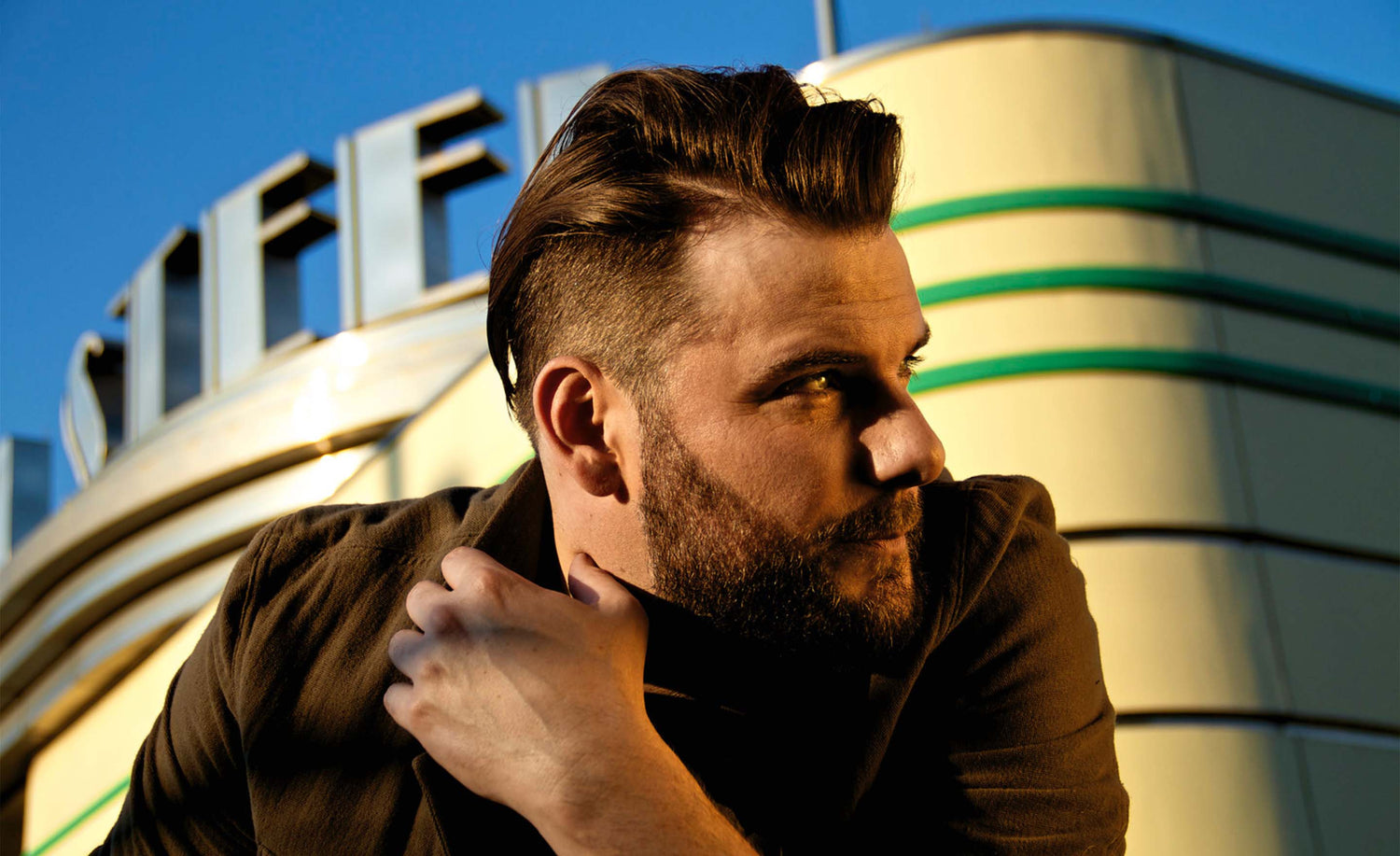 Male musician portrait Brad James hand up against shoulder looking far left with steel factory behind